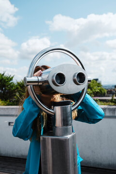 Anonymous blond female in blue coat looking through street coin operated binocular while standing on wooden pavement near concrete fence on sunny day