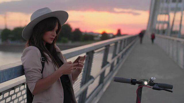 A young smiling woman is leaning on the bridge fence and typing on her phone. An electric scooter is parked next to her. River, sunset and the parts of a modern bridge predominate in the background.
