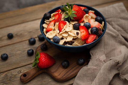 From above delicious breakfast bowl of corn flakes with strawberries and blueberries placed on cutting board and decorated with linen cloth and berries around dish on wooden background