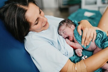 High angle of cheerful adult woman embracing newborn child covered with blood after giving birth in delivery room of contemporary hospital