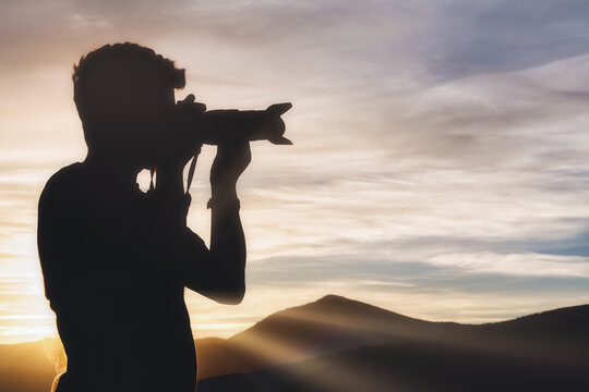 Side view of young male traveler in silhouette standing on hill and taking picture of spectacular scenery of mountain range on sunset