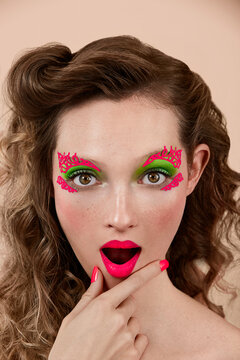 Astonished young lady with colorful makeup rubbing chin and looking at camera with open mouth against beige background