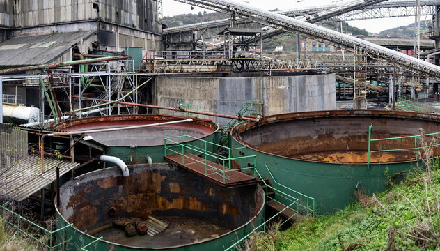 From above of huge rusted metal empty water tanks painted green surrounded by various dilapidated structures and trolleys for transporting coal in idle abandoned coal mine