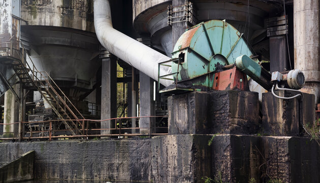 Part of huge steel rusty mechanism standing on dirty brick black blocks next to large cylindrical structures in abandoned desert plant