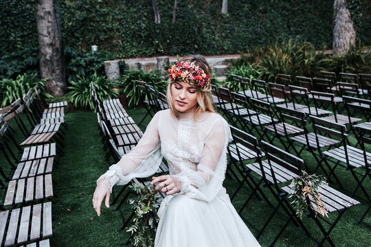 High Angle Of Beautiful Young Female In Elegant White Dress And Flower Wreath Closing Eyes And Sitting On Empty Chairs Before Wedding Ceremony In Garden