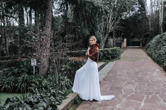 Side view of young lady in white dress and short fur coat looking at camera while stepping on border of weathered path during wedding in green garden