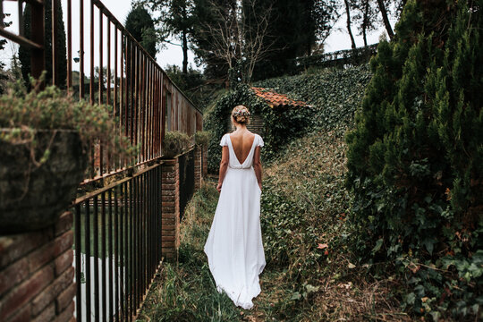 Back view of unrecognizable female in elegant white dress walking near metal fence on wedding day in green garden