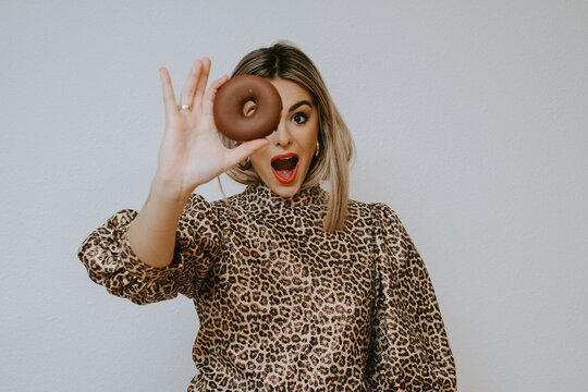 Young cute blonde female in dress with leopard print pouting lips and looking at camera through sweet chocolate doughnut against gray background