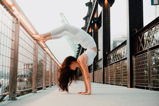 Low angle side view of flexible female in sports bra and leggings performing scorpion handstand while exercising barefoot and leaning on metal railing in city