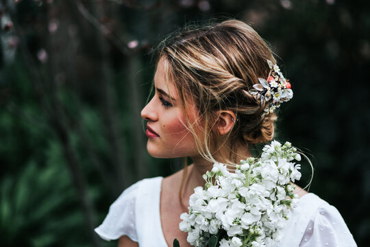 Pretty blond woman with bunch of white flowers looking away while standing on blurred background of garden during wedding on summer day