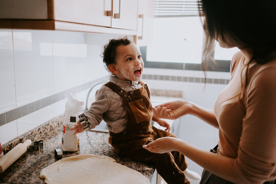 Side view of cheerful little boy in casual clothes with flour on cheeks helping mother cooking pastry in modern kitchen