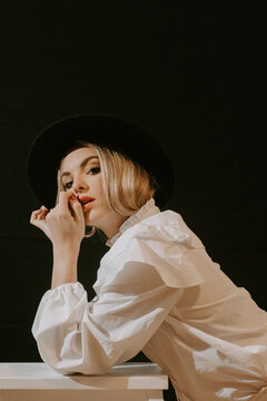 Side view of young blonde woman in white blouse and stylish hat looking at camera while leaning on stool against black background