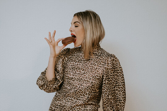 Happy young blonde female in trendy dress with leopard print smiling with closed eyes biting tasty chocolate doughnut while standing against gray wall background