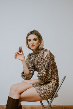 Happy young blonde female in trendy dress with leopard print smiling looking at camera biting tasty chocolate doughnut while sitting on chair against gray wall background