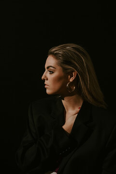 Side view of young blonde woman in stylish black jacket looking away against black background