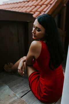 High angle of young barefoot woman in trendy red dress sitting lumber floor while resting on terrace