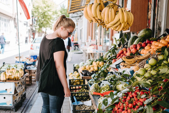 Side view of positive female in casual wear and sunglasses standing near fruit counter in Turkish market and exploring goods while walking through streets of city