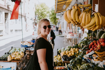 Side view of positive female in casual wear and sunglasses looking at camera standing near fruit counter in Turkish market and exploring goods while walking through streets of city