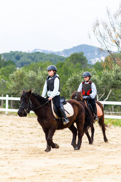 Teen jockeys in helmets riding obedient horses on sandy dressage arena during lesson in equestrian school