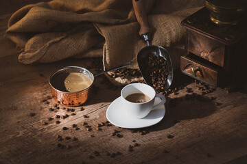 Fresh black coffee in white ceramic cup placed on saucer near coffee grinder and coffee beans on wooden table
