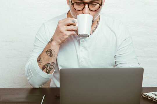 Focused male in glasses with tattoos on arms drinking coffee and surfing laptop while working at home because of quarantine