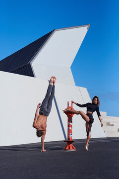 Upside down shirtless male acrobat in jeans doing handstand with one hand while graceful partner in pointe shoes performing element of ballroom dance standing on tiptoe with one leg raised on rooftop of modern building