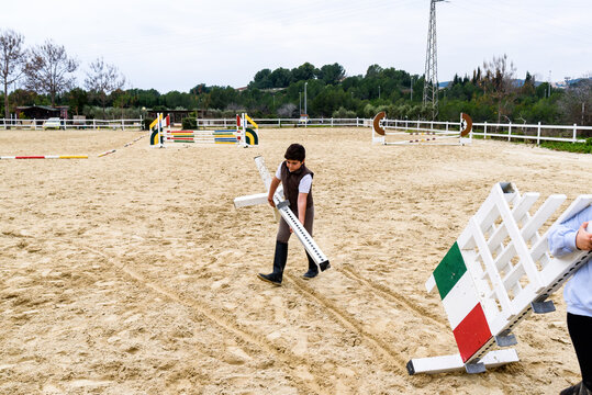 Little jockey carrying hurdle parts while disassembling dressage equipment on arena after training in equestrian school