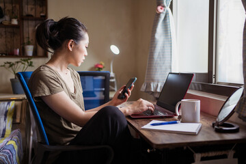 Side view of Asian female freelancer in casual t shirt sitting at table and browsing computer working on project online at home while texting on the smartphone