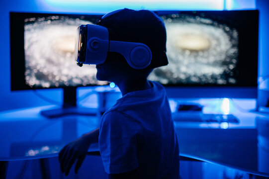 Side view of anonymous male kid in t shirt and VR glasses watching at desktop computer screen while sitting in room with blue light.
