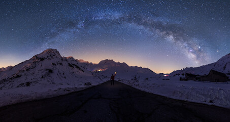 Breathtaking view of distant silhouette of explorer standing on road leading to snowy mountainous area and holding luminous light in hand against starry stunningly beautiful sky on winter night