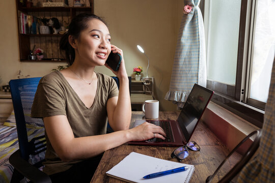 Side view of Asian female freelancer in casual t shirt sitting at table and browsing computer working on project online at home while speaking o the smartphone