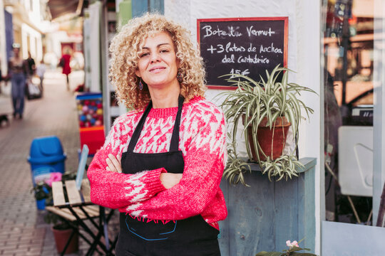 Positive adult woman with crossed arms smiling and looking at camera while standing near entrance of local food store
