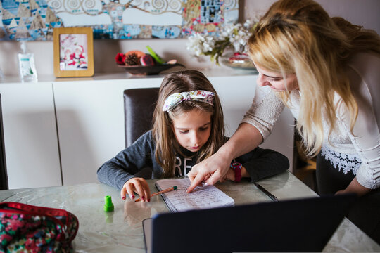 Adult woman pointing at task in notebook while helping girl to do homework in cozy room at home