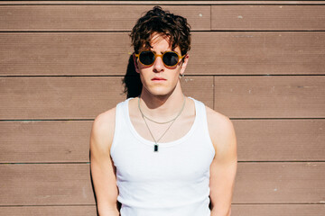 Modern young handsome man in trendy sunglasses and white tank top standing near brown wall on sunny day on city street