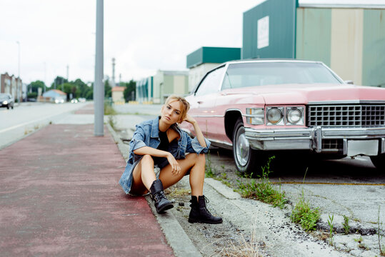Attractive blonde girl sitting on the sidewalk near to a classic pink car