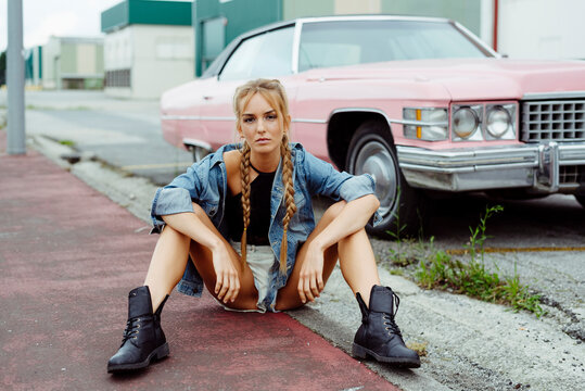 Attractive blonde girl sitting on the sidewalk near to a classic pink car
