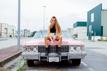 Attractive blonde girl sitting on the hood of a classic pink car on the street
