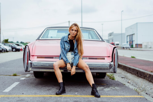 Sensual blonde girl sitting on the bumper of a classic pink car looking at  the camera Stock Photo | Adobe Stock