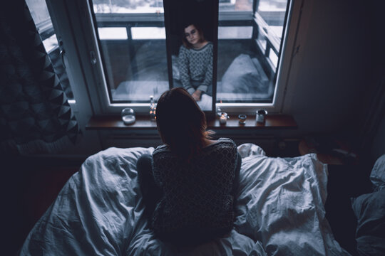 Back view of depressed young female sitting on bed in dark bedroom looking at mirror at home