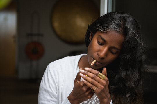 Young Indian female in casual clothing standing with closed eyes on terrace and smoking marijuana roll up while relaxing