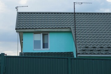 part of the house with one white window on a blue wall under a green tiled roof behind a metal fence against a gray sky and clouds