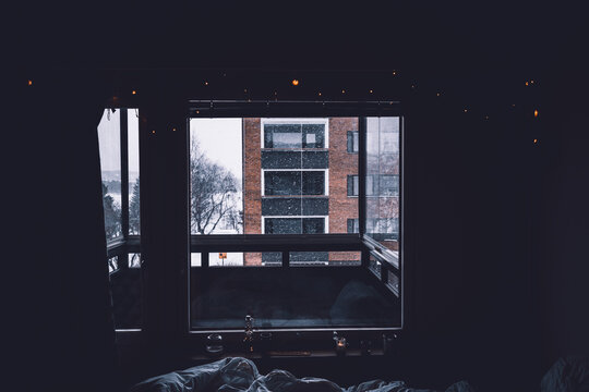 Apartment building located on snowy street behind balcony and window of cozy dark room