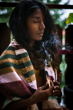 Spiritual young ethnic female with closed eyes in traditional clothes holding burning sage incense with smoke and meditating outdoors