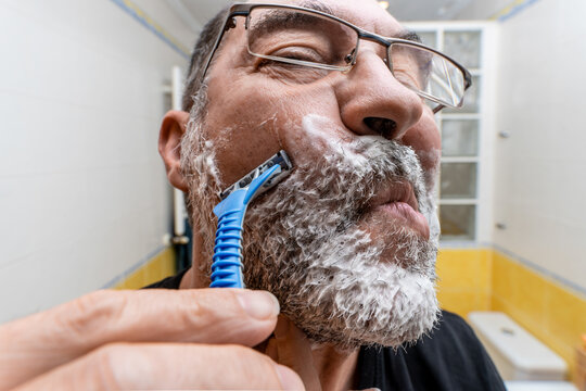 Low angle closeup of middle aged bearded male shaving with disposable razor in front of mirror in bathroom