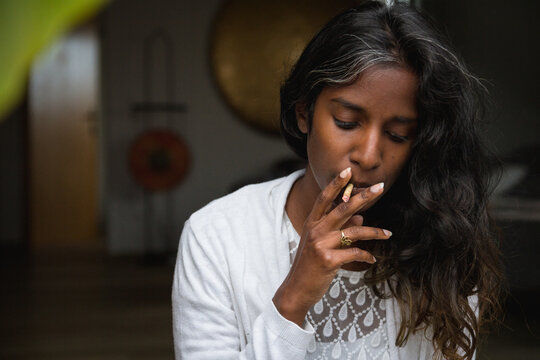 Young Indian female in casual clothing standing with closed eyes on terrace and smoking marijuana roll up while relaxing