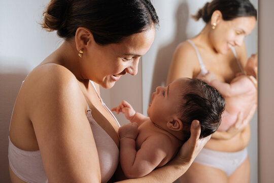 Delighted woman in underwear cuddling naked baby near mirror while leaning on wall and smiling