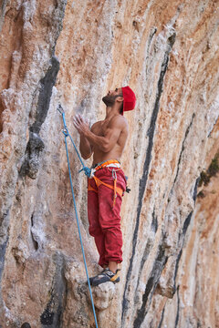 Side view of muscular shirtless bearded alpinist in red cap and safety equipment standing near rocky slope of mountain and looking up with hands together
