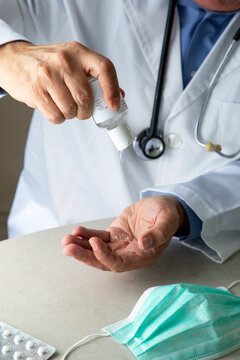 Male therapist in medical gown sitting at table in hospital and sanitizing hands with antiseptic while preparing for treatment of patients during coronavirus outbreak