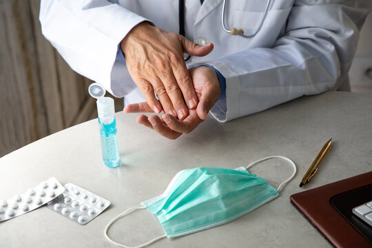 Male therapist in medical gown sitting at table in hospital and sanitizing hands with antiseptic while preparing for treatment of patients during coronavirus outbreak