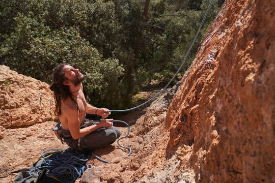 From above view of active bearded shirtless male climber sitting on edge of cliff and holding rope in hands while looking up